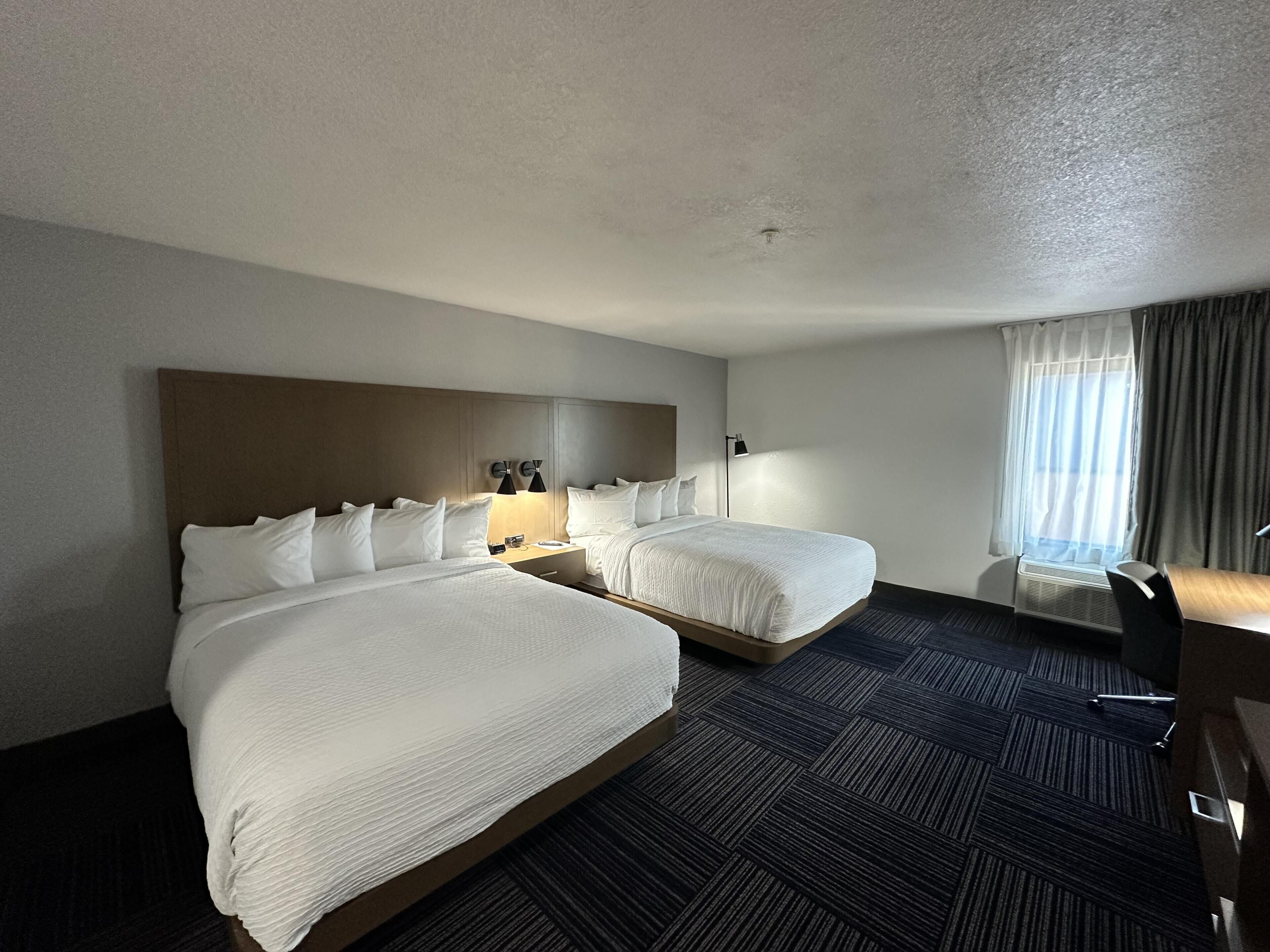 Habitación de hotel con dos camas, un escritorio, una silla, una mesita de noche y una ventana con cortinas.