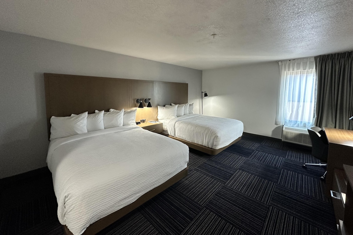 A hotel room with two beds, a desk, a chair, a nightstand, and a window with curtains.