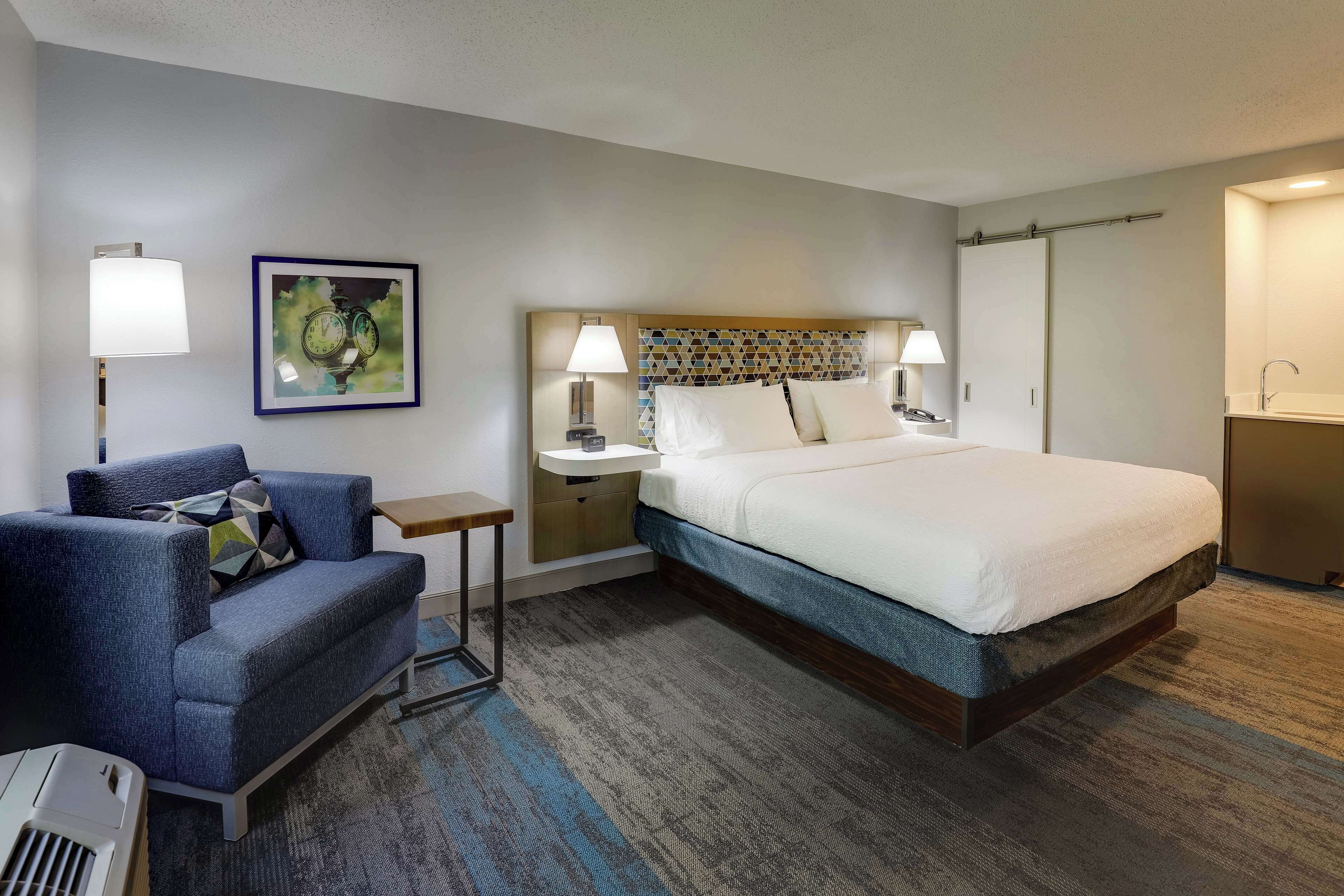 A hotel room with a bed, a desk, a chair, a lamp, and a painting on the wall.