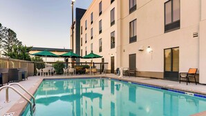 A swimming pool with a tiled edge, surrounded by outdoor seating with umbrellas, and a building with multiple windows and a door.