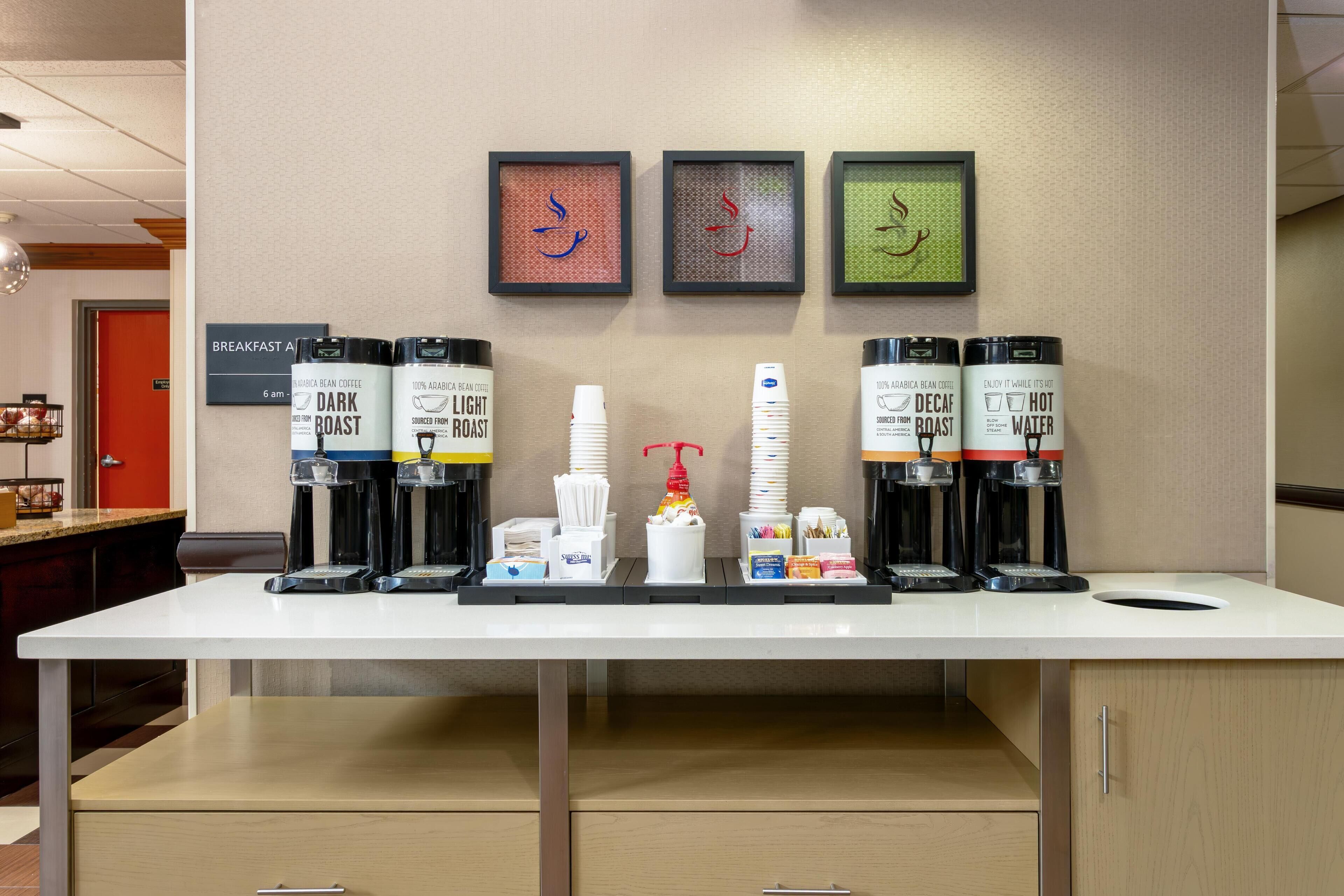 A coffee station with multiple coffee dispensers, a coffee machine, and a sign indicating breakfast hours.