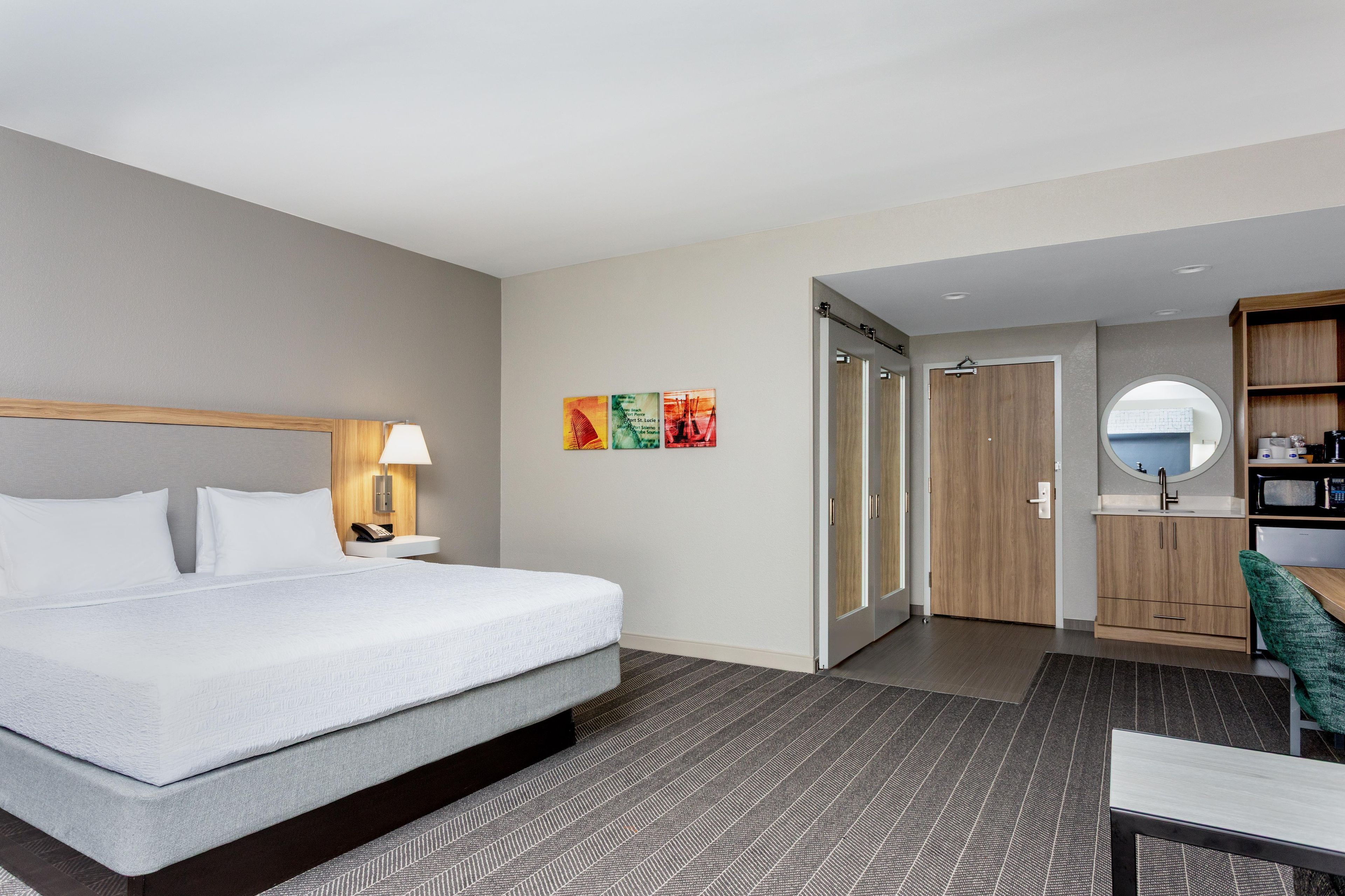 Ein Hotelzimmer mit Bett, Nachttischlampe, Kunstwerk, einem Badezimmer mit Spiegel und einer Küchenzeile.