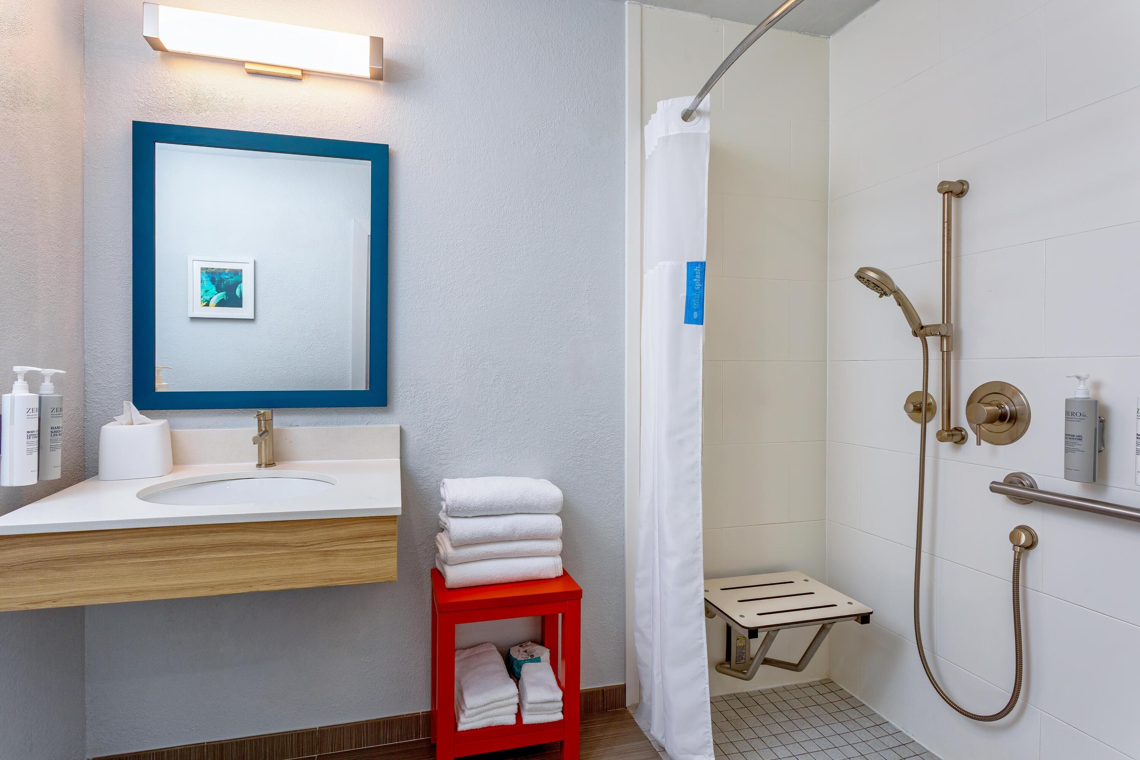 A bathroom with a sink, mirror, towel rack, shower, and a small shelf.