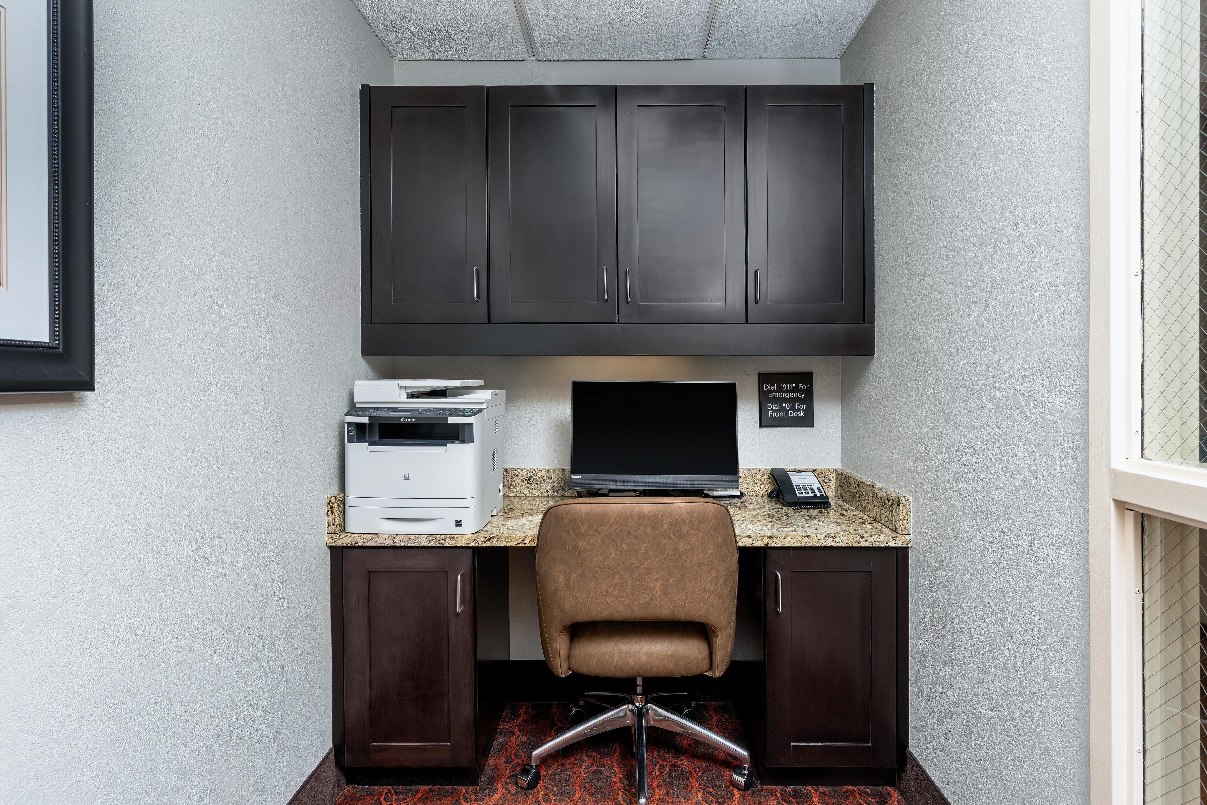 A corner office with a dark wood desk, a brown leather chair, a printer, a monitor, and a phone.