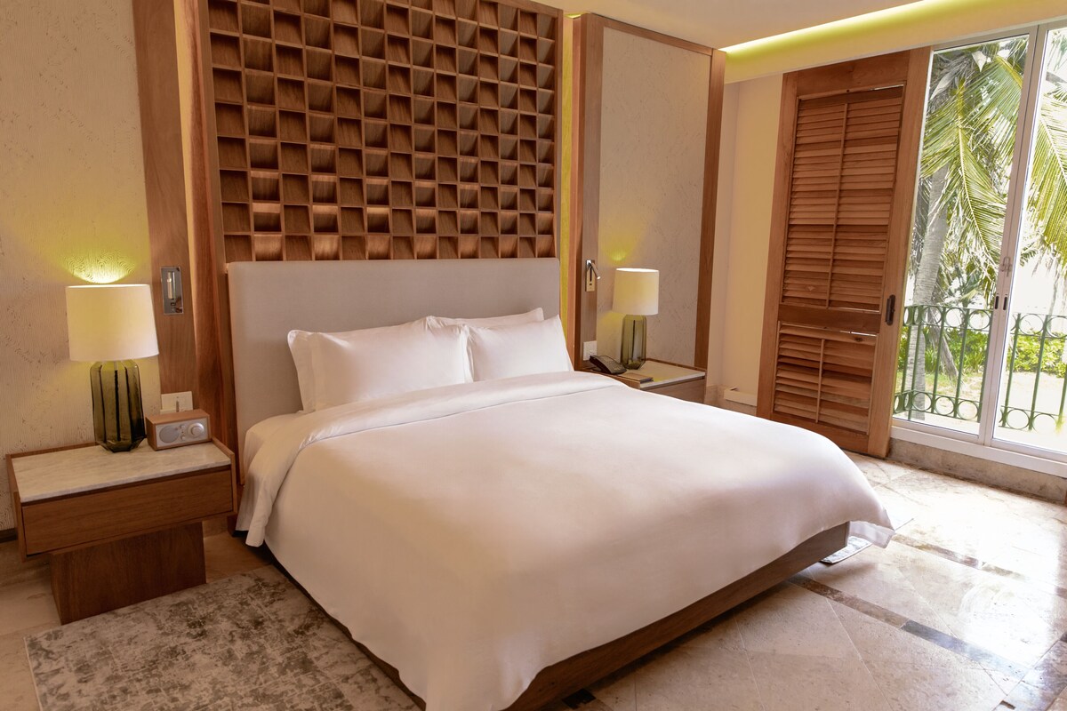 A hotel room with a large bed, wooden headboard, two bedside tables with lamps, a small table with a radio, and a view of palm trees through a sliding door.