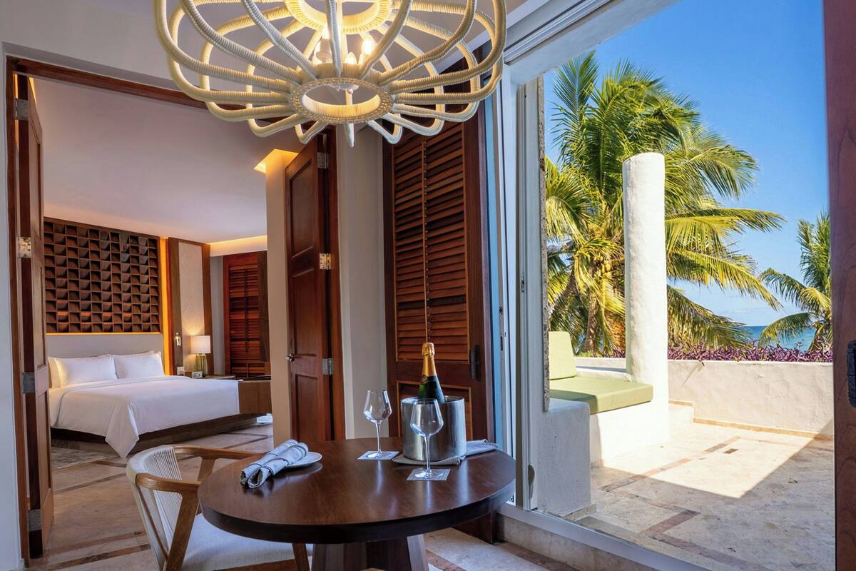Habitación de hotel con cama, mesita de noche y vista a palmeras y al océano.