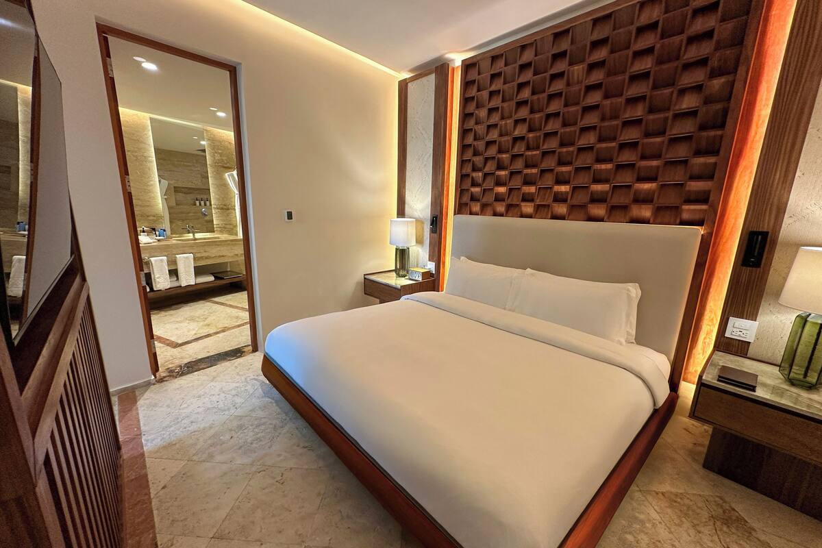 A hotel room with a large bed, a wooden headboard, a nightstand with a lamp, and a bathroom visible through an open door.
