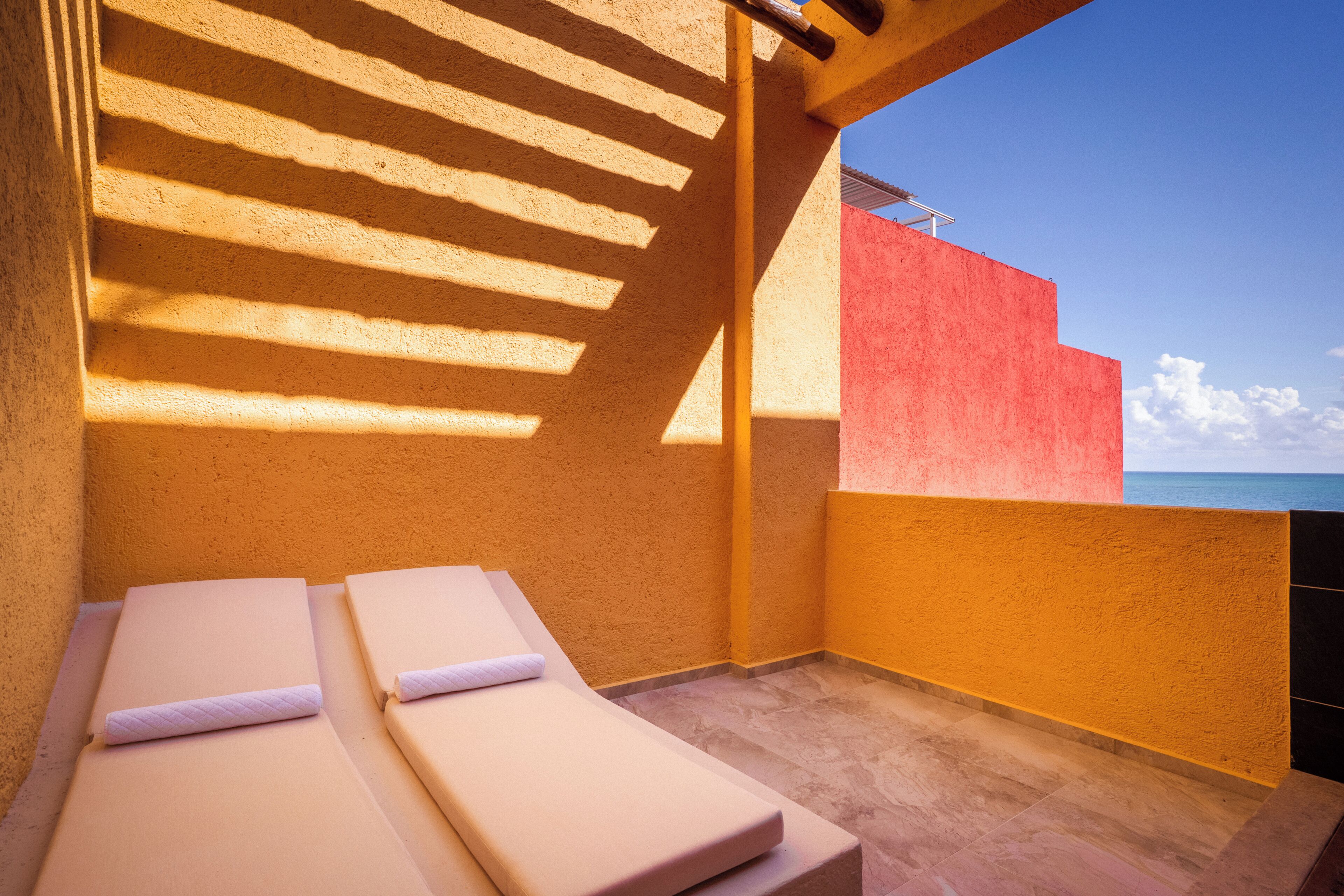 Un balcón soleado con dos sillones de descanso, una pared roja y vista al mar.