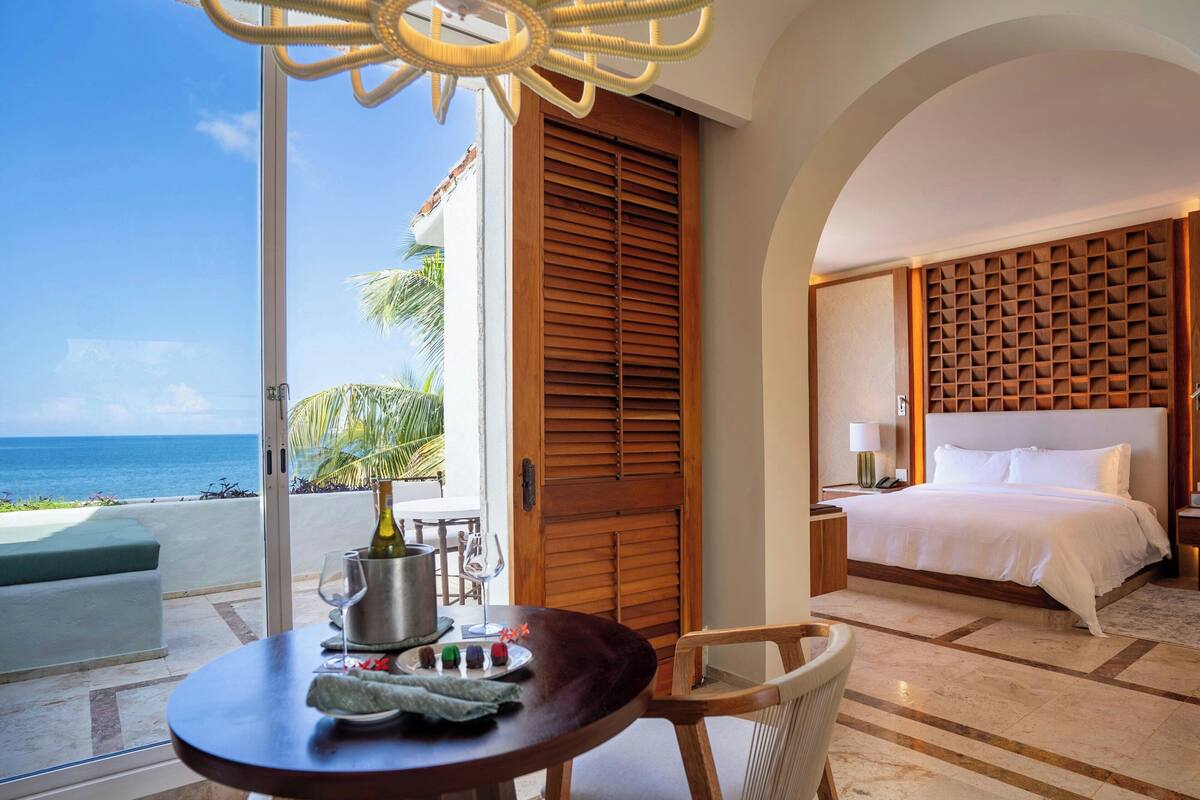 Habitación de hotel con cama, una mesa de comedor con una botella y vasos, vista al océano y una puerta de madera.