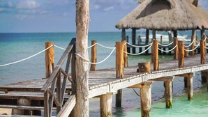Un muelle de madera que lleva a una edificación de techo de paja sobre aguas turquesas.