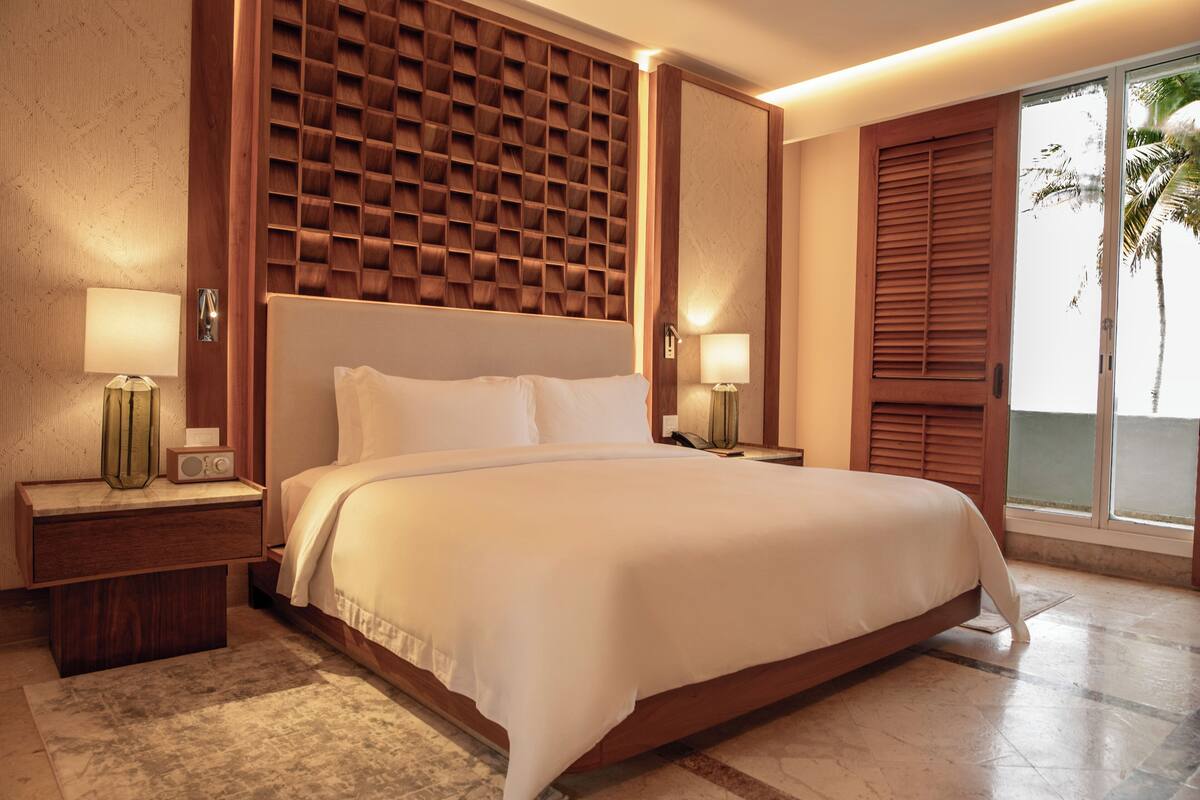 A hotel room with a large bed, wooden headboard, two bedside tables with lamps, a large window with a view of palm trees, and a sliding door.