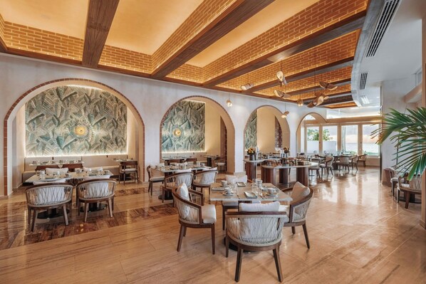 A spacious dining area with high ceilings, brick accents, and a central mural. Tables are neatly arranged with chairs, and the floor is polished wood.
