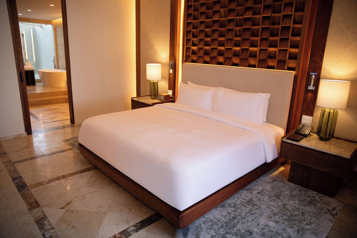 A hotel room with a large bed, wooden headboard, two bedside tables with lamps, and a bathroom visible through an open door.