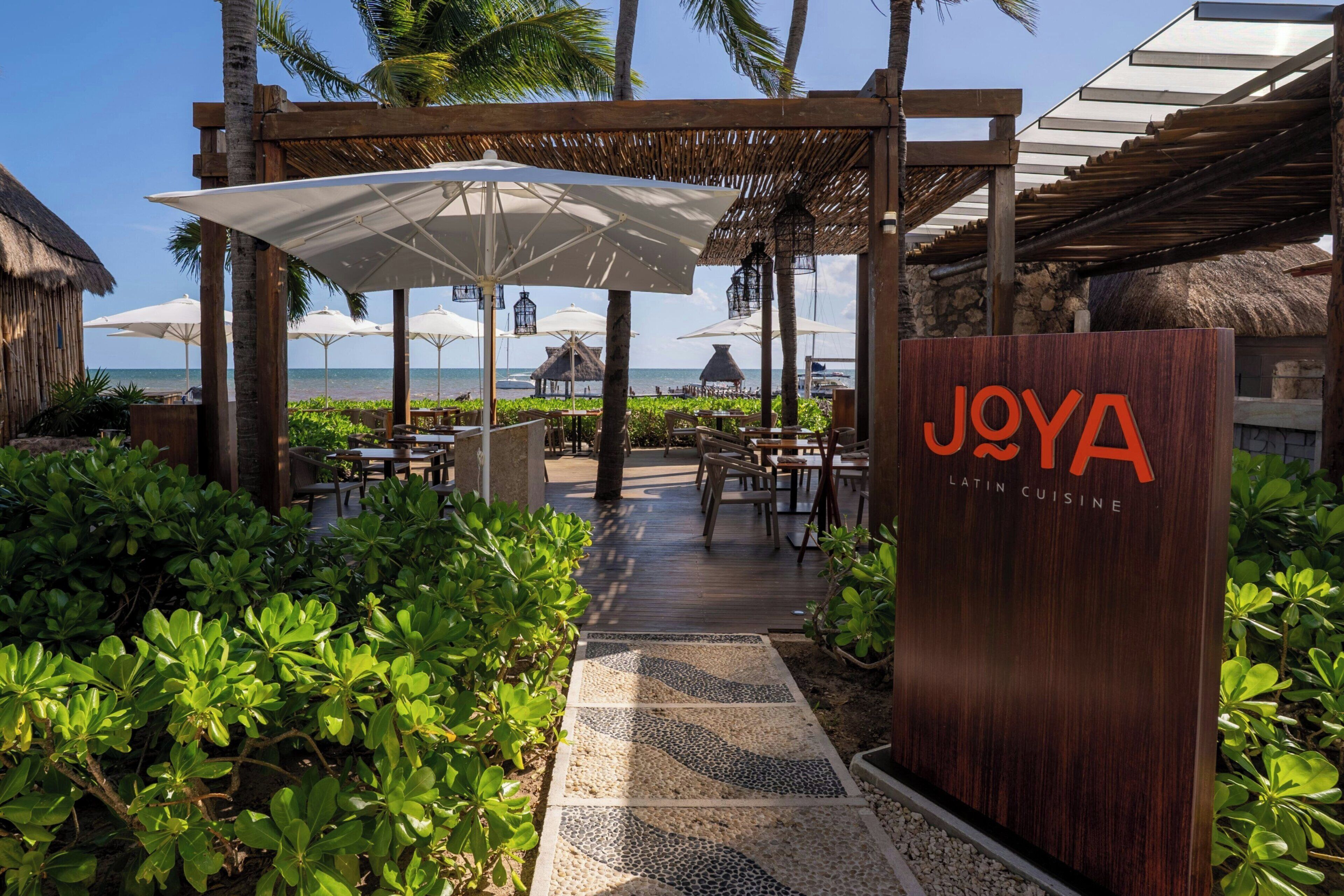 A wooden sign with 'JOYA LATIN CUISINE' and a pathway leading to outdoor seating with umbrellas.