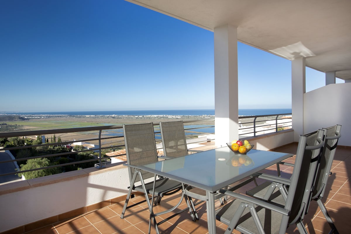 En balkon med et glasbord og stole, en skål med frugt og udsigt over havet og det fjerne landskab.
