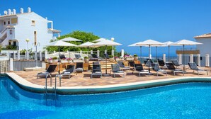 Ein Poolbereich mit Liegestühlen und Sonnenschirmen, eine geflieste Terrasse und ein Gebäude mit weißer Fassade und roten Ziegeldach.
