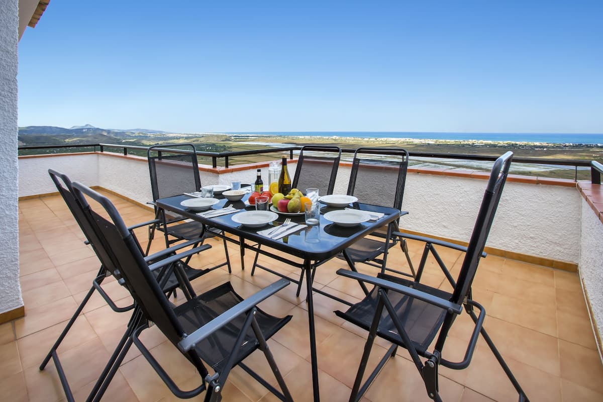 Et spisebord på en balkon med stole, tallerkener, bestik og en vinflaske, med udsigt over et kystlandskab.
