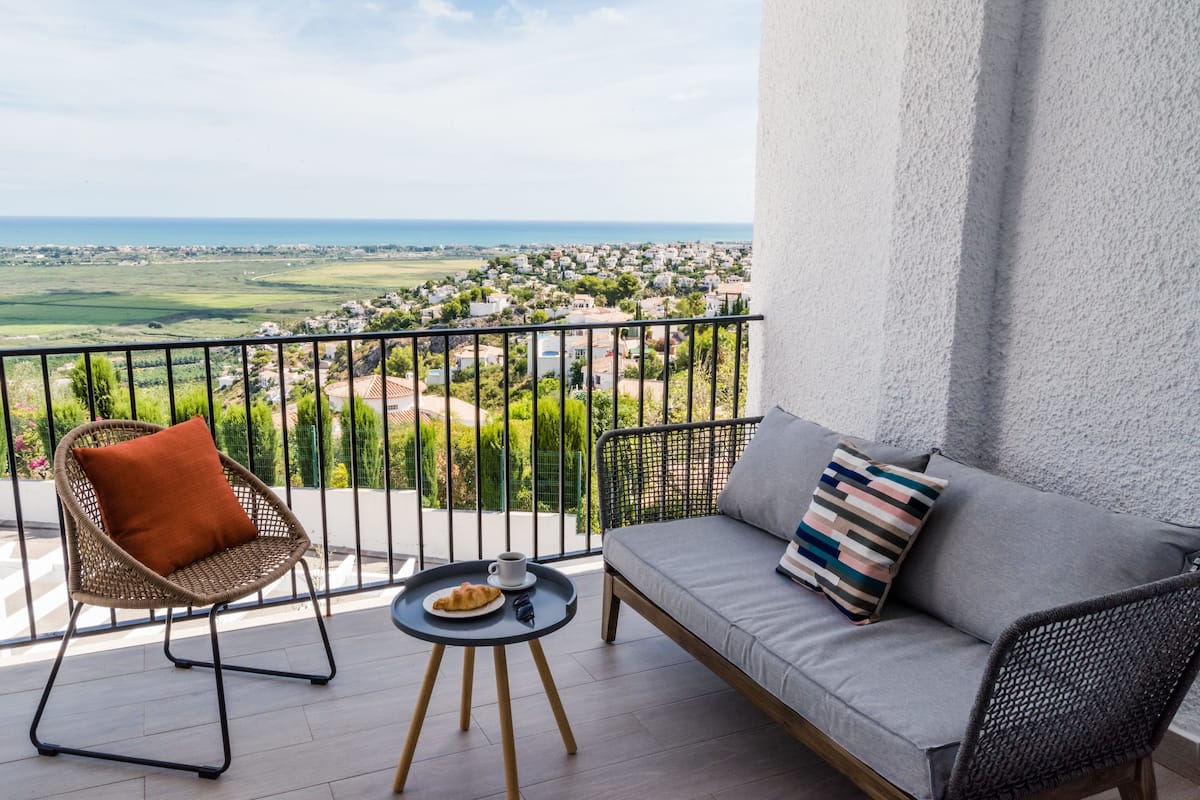 En balkon med en sofa, en fletstol, et lille rundt bord med mad og en kop, og udsigt over en kystby.