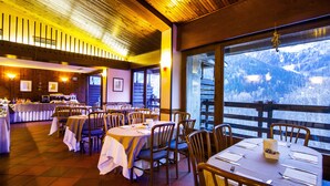 A restaurant with multiple tables set for dining, wooden chairs, and a view of a mountainous landscape through large windows.