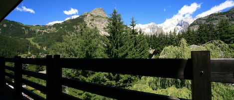 A wooden railing overlooking a mountainous landscape with dense forests and snow-capped peaks under a clear blue sky.