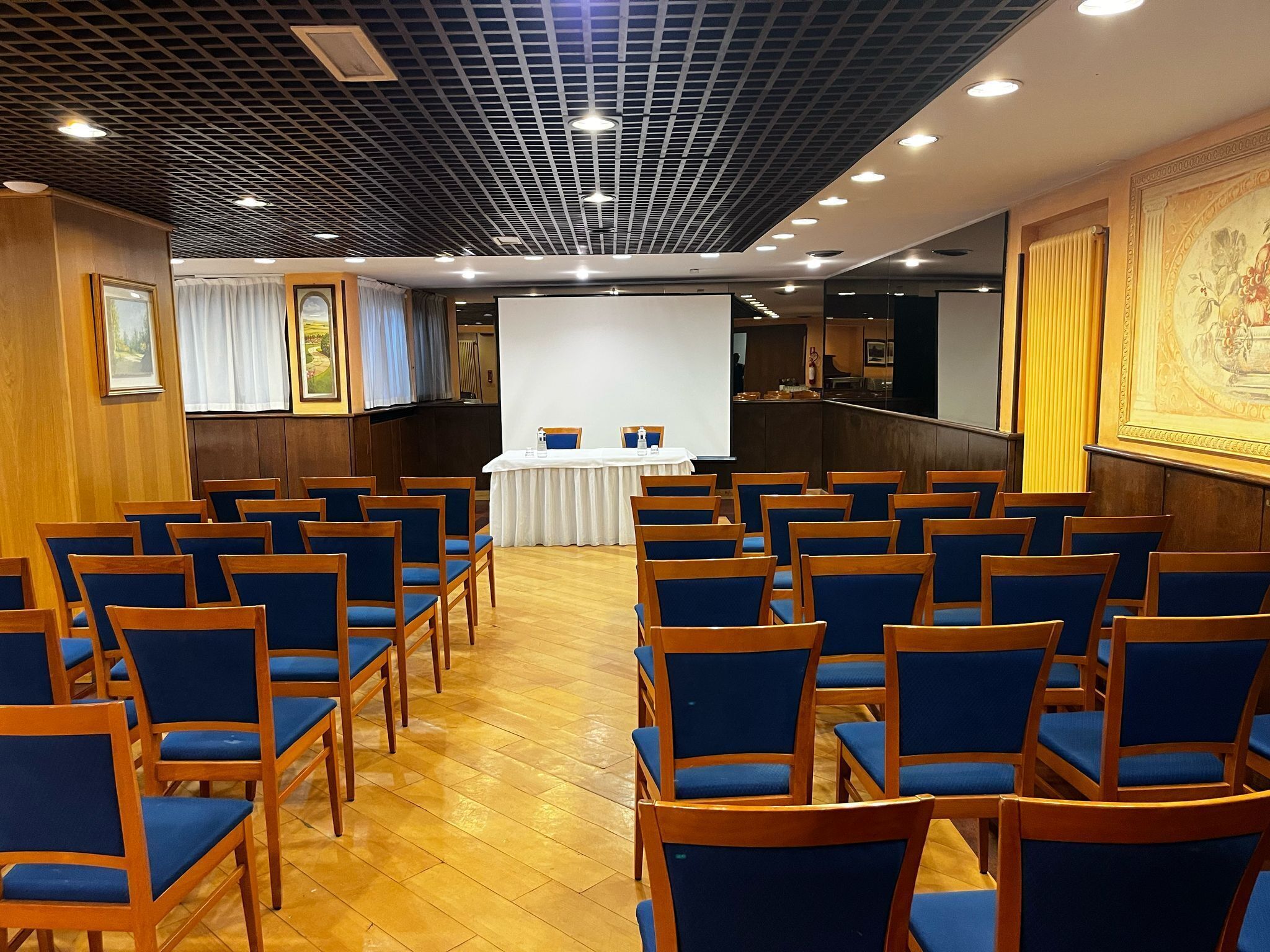 Une salle de conférence avec des rangées de chaises bleues, une table blanche avec deux chaises et un écran de projection.