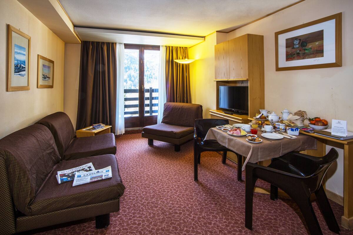 Une chambre d’hôtel avec un canapé, une petite table avec une lampe, une table à manger dressée avec des assiettes et des tasses, et un téléviseur posé sur un meuble.