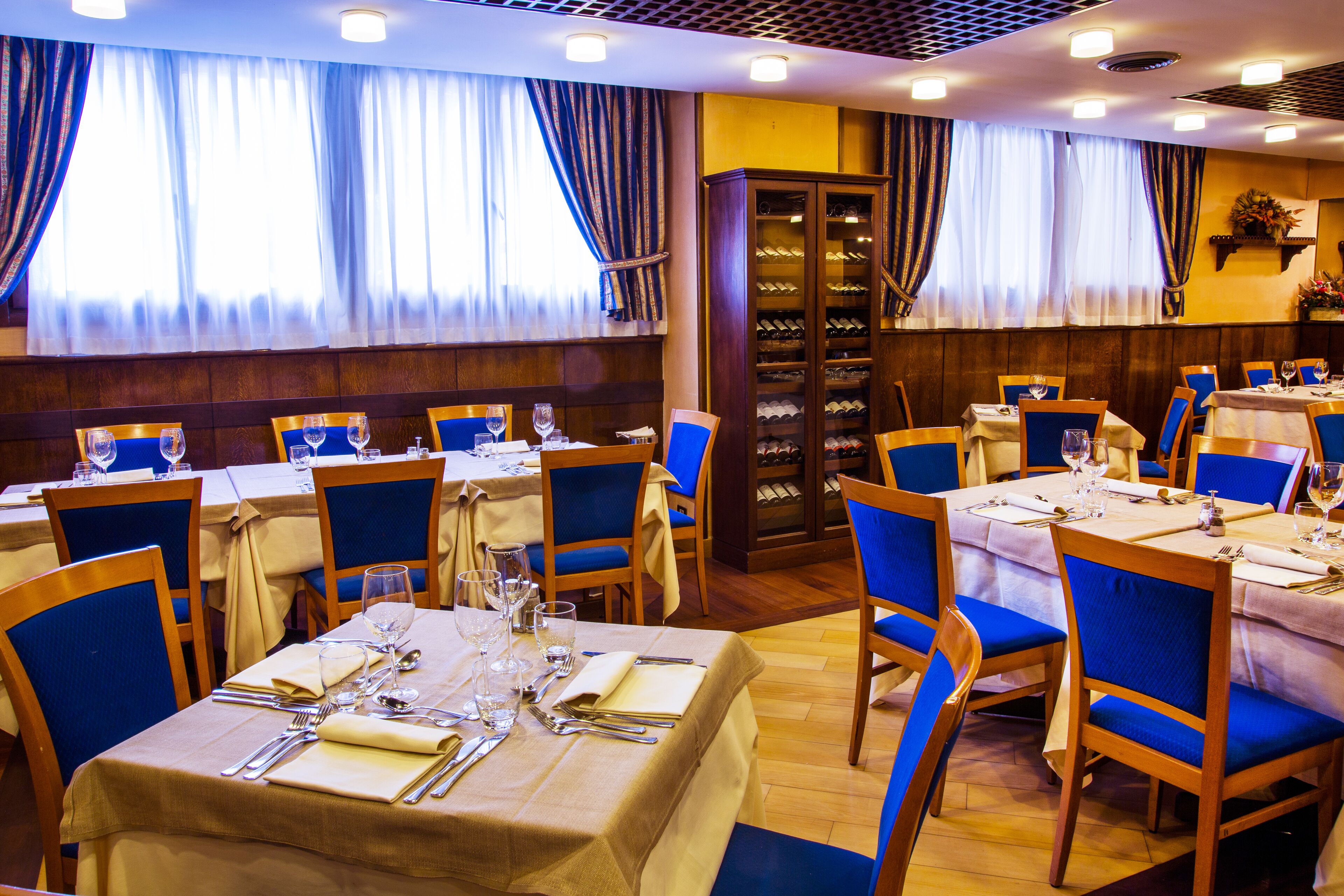 A restaurant with multiple tables set for dining, blue chairs, and a wine cabinet.