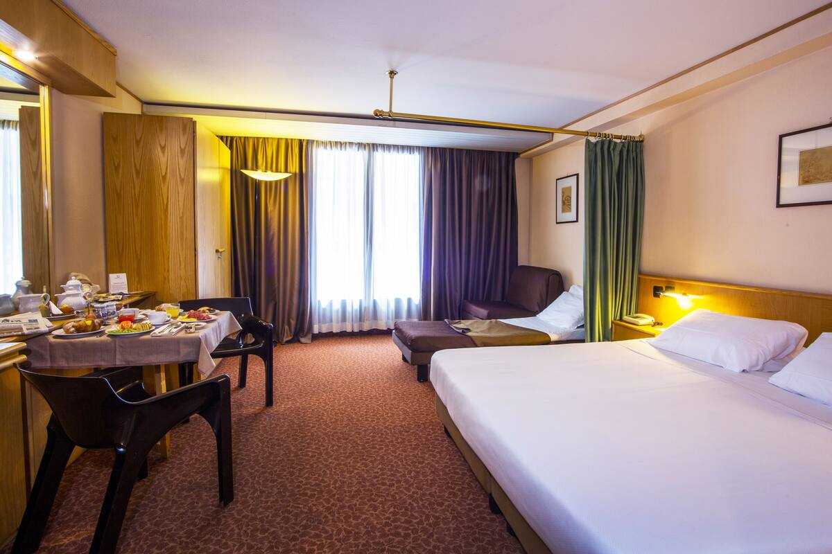 Une chambre d’hôtel avec un lit, un bureau avec un petit-déjeuner, une chaise et une fenêtre avec des rideaux.