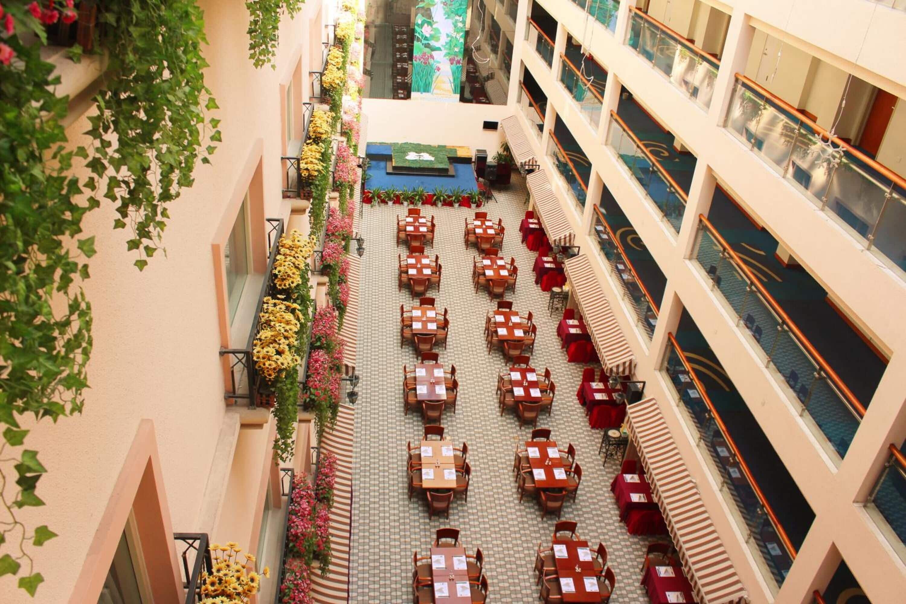 A multi-level hotel atrium with rows of tables and chairs, indoor plants, and a central decorative area.
