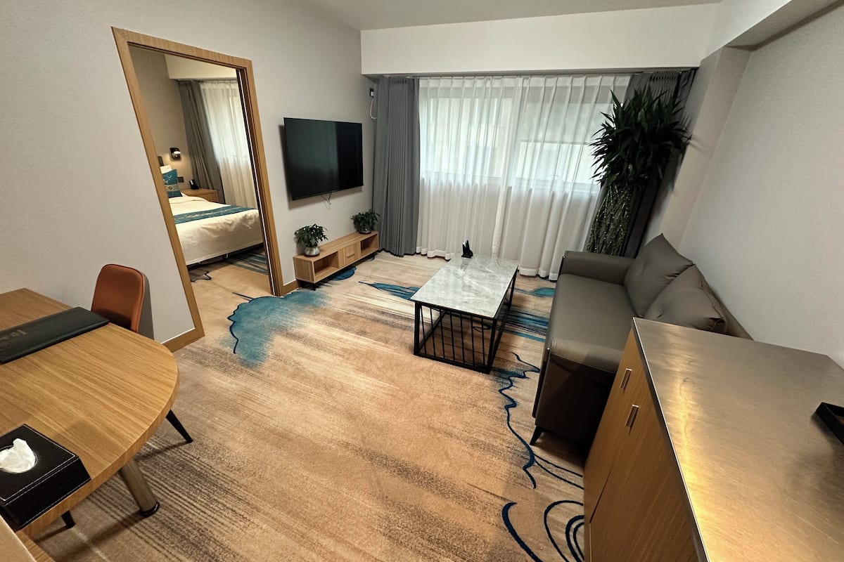 A modern hotel room with a large bed, a TV, a sofa, a dining table, and a mirror.