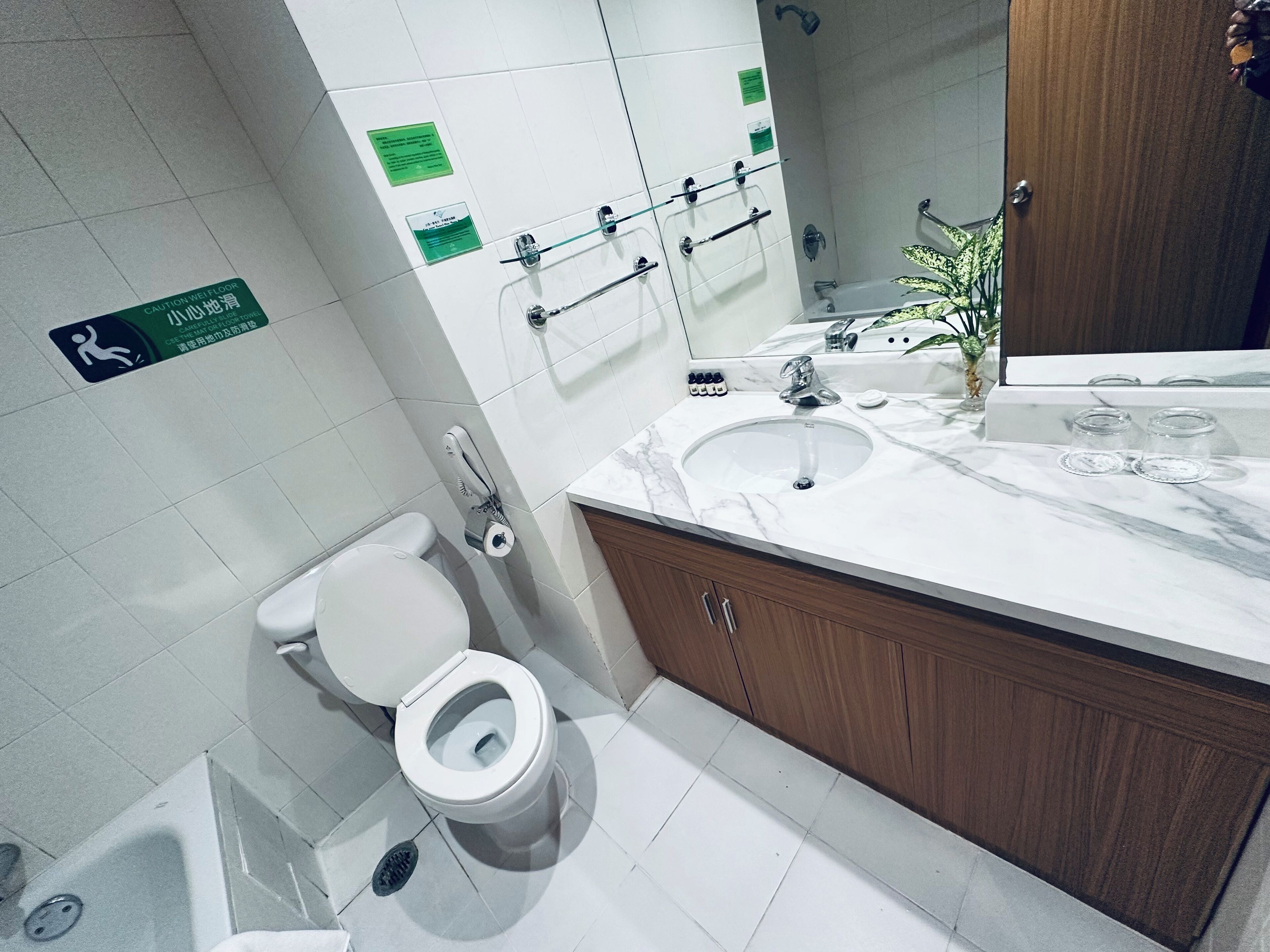 A modern bathroom with a white toilet, a sink with a marble countertop, and a mirror above the sink. There are multiple towel hooks on the wall and a green caution sign on the floor.