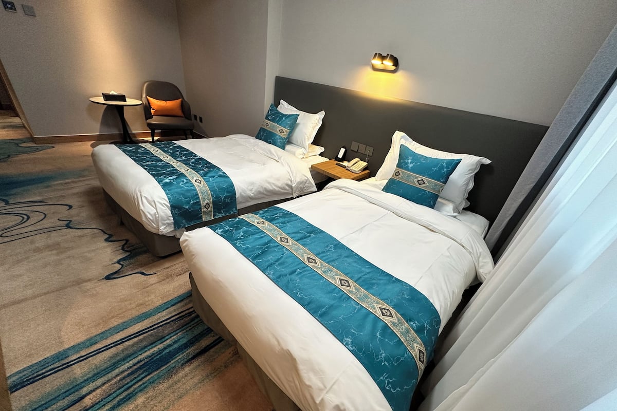 A hotel room with two beds, each with a blue and white bedspread, a small table with a chair, a wall-mounted light, and a telephone on the nightstand.