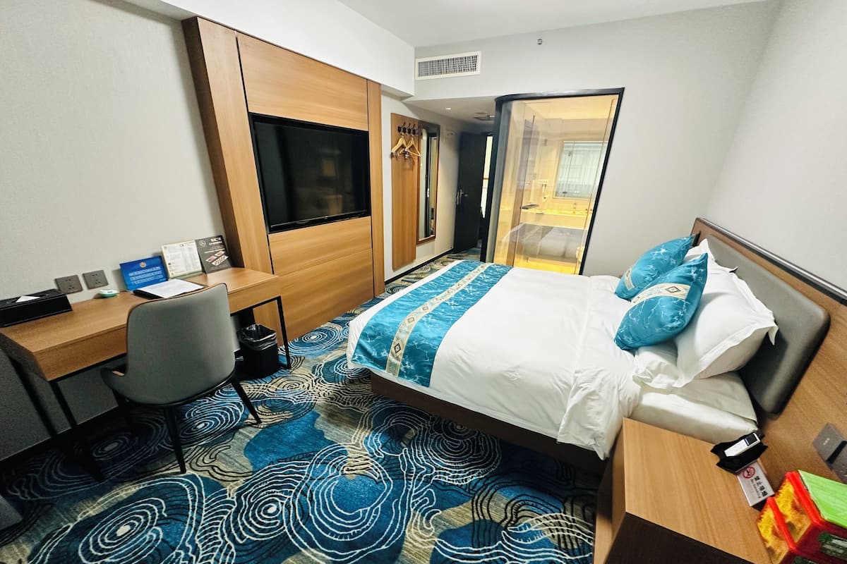 A hotel room with a bed, a desk, a chair, a TV, and a bathroom visible through a glass door.