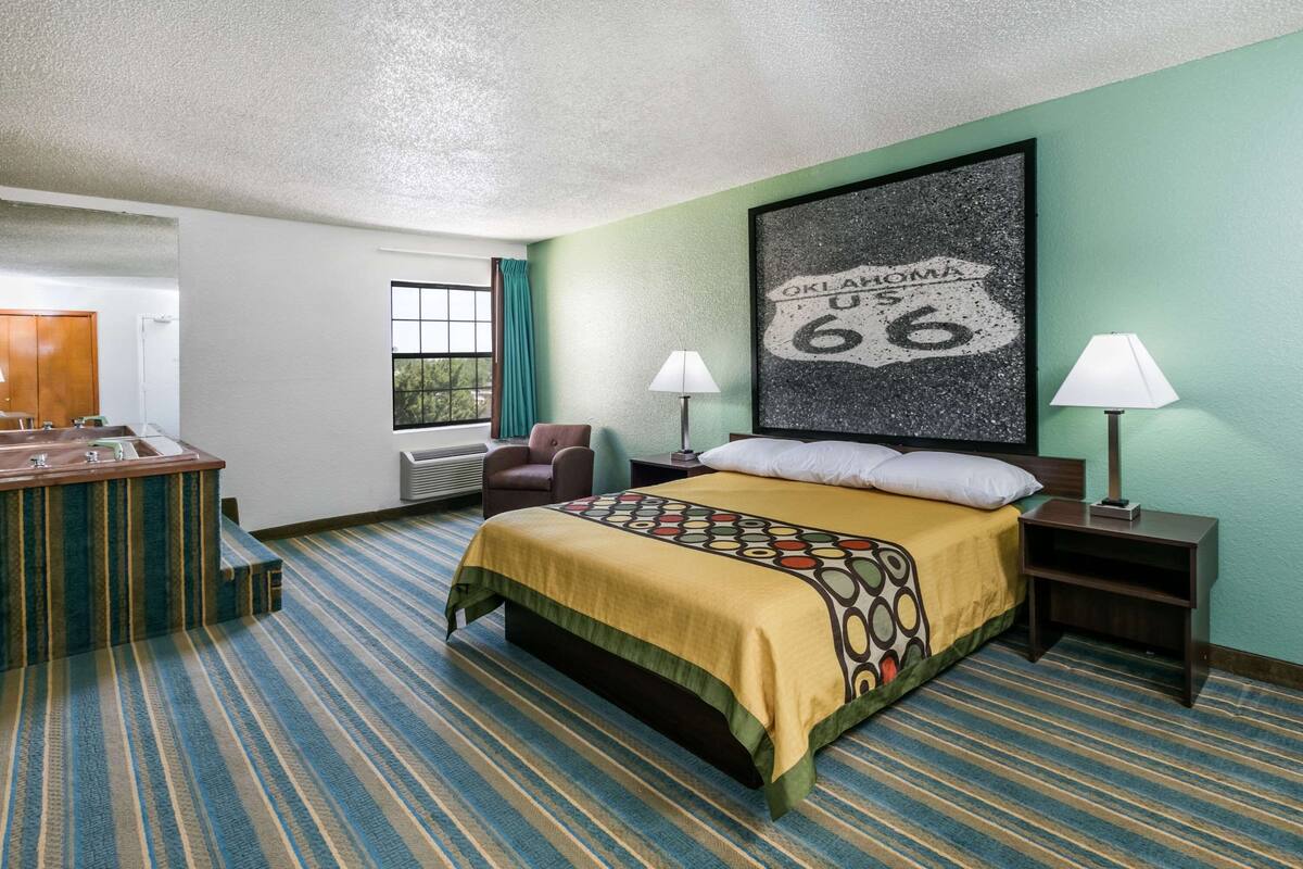 A hotel room with a bed, two bedside tables with lamps, a desk with a chair, a window with curtains, and a wall with a framed picture.