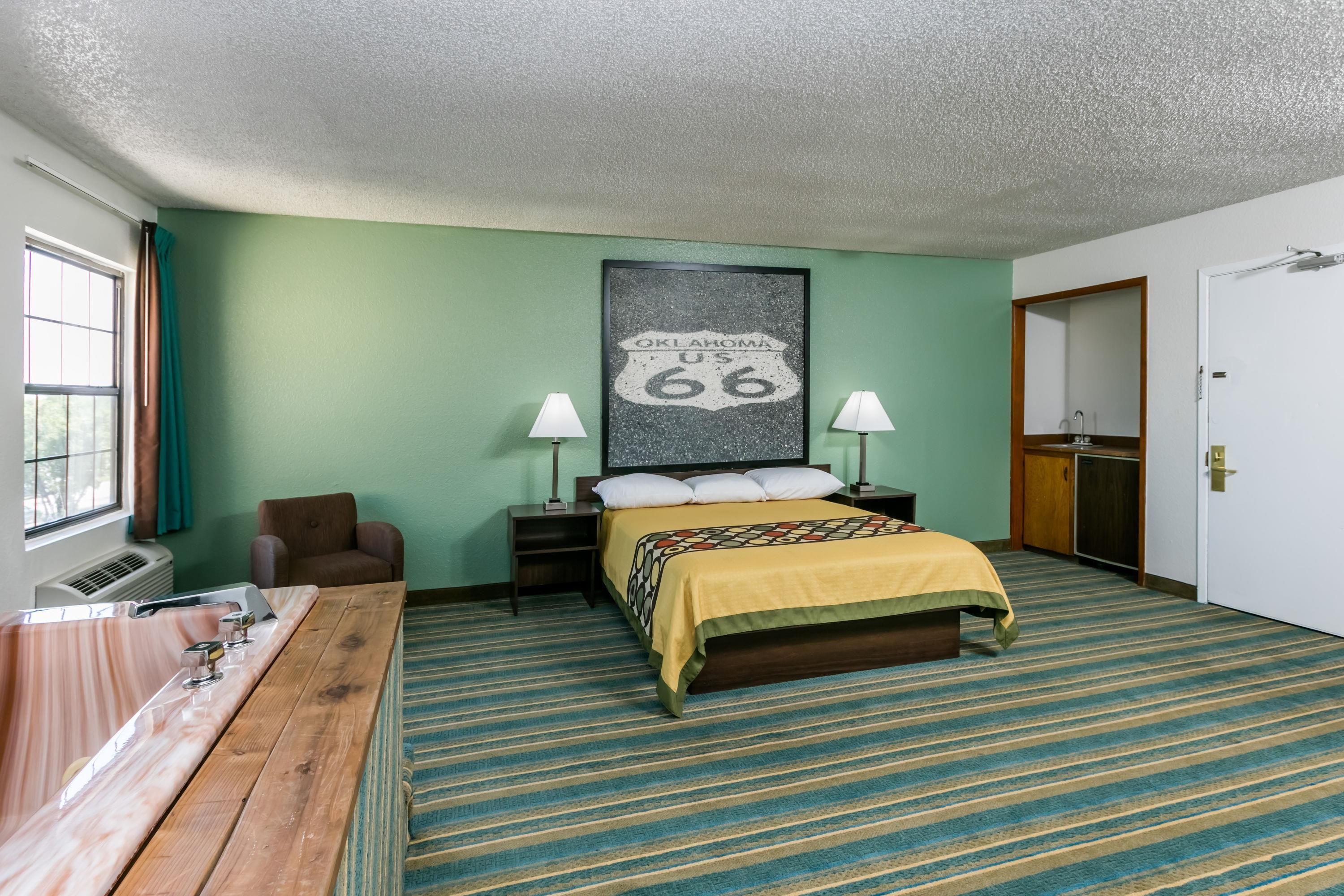 A hotel room with a bed, a desk with a sink, a chair, a window with curtains, and a framed picture on the wall.