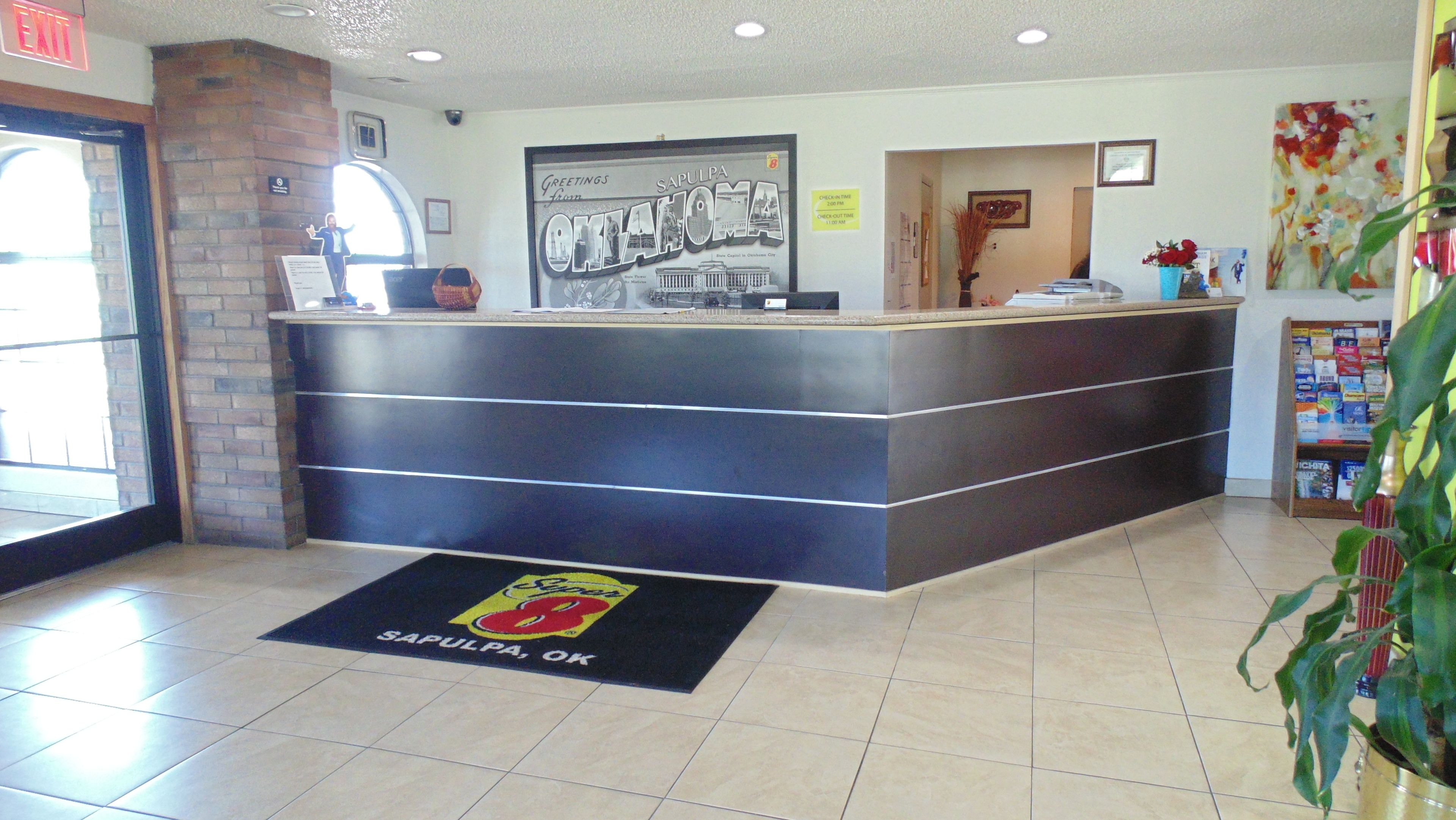 Una recepción con un cartel de bienvenida enmarcado, un letrero con el horario de registro y un felpudo con el logotipo del hotel.