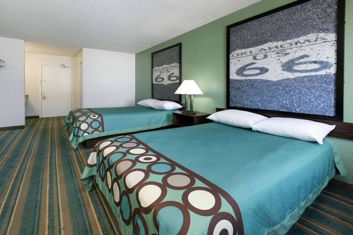 A hotel room with two beds, a nightstand, a lamp, and a large window with a view of a road sign.