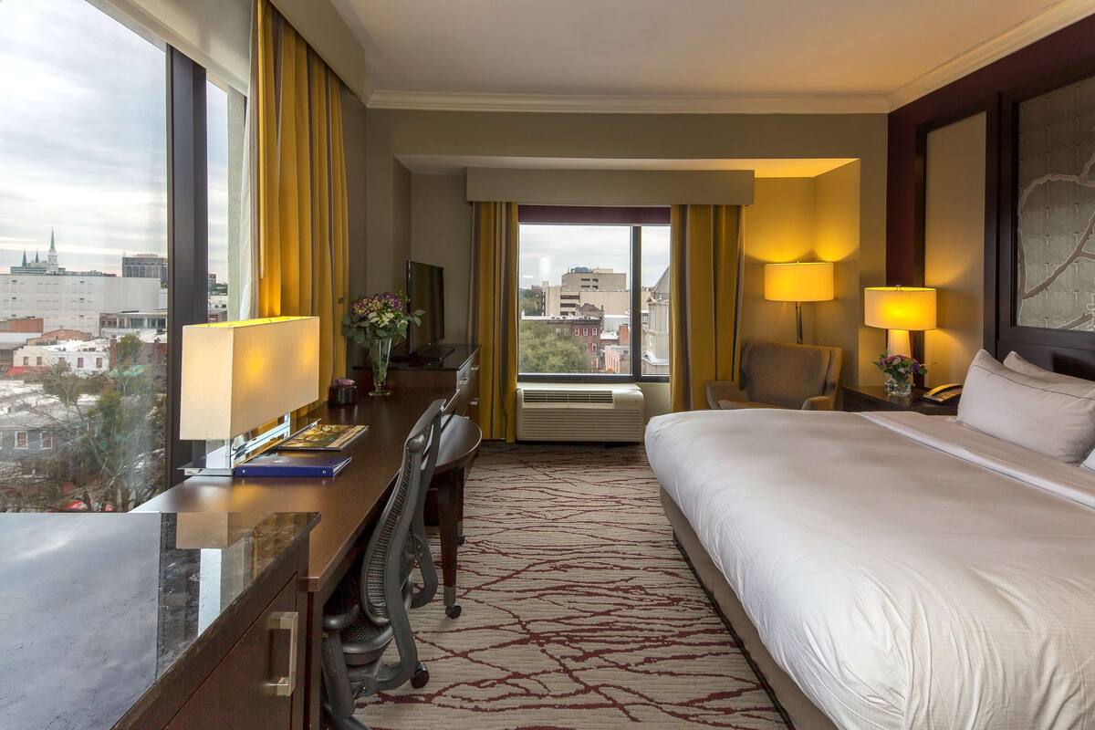 A hotel room with a large bed, a desk with a computer, a chair, and a view of the cityscape through large windows.