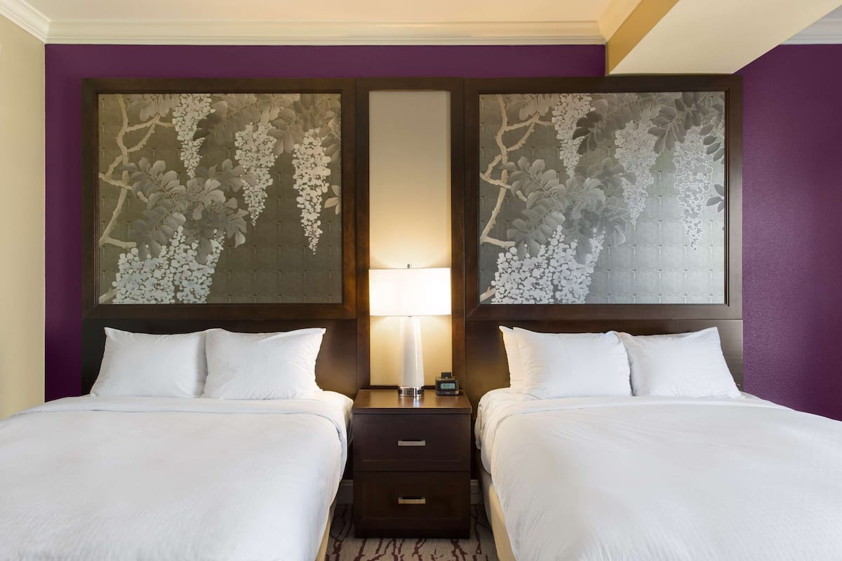 A hotel room with two beds, a nightstand with a lamp, and a large floral artwork on the wall.