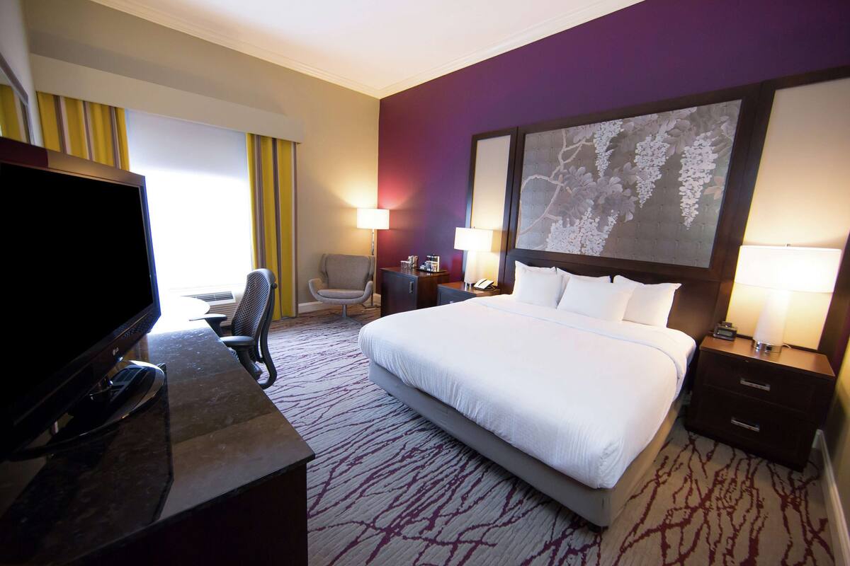 A hotel room with a large bed, a desk with a flat-screen TV, two bedside lamps, a chair, and a large wall art piece.