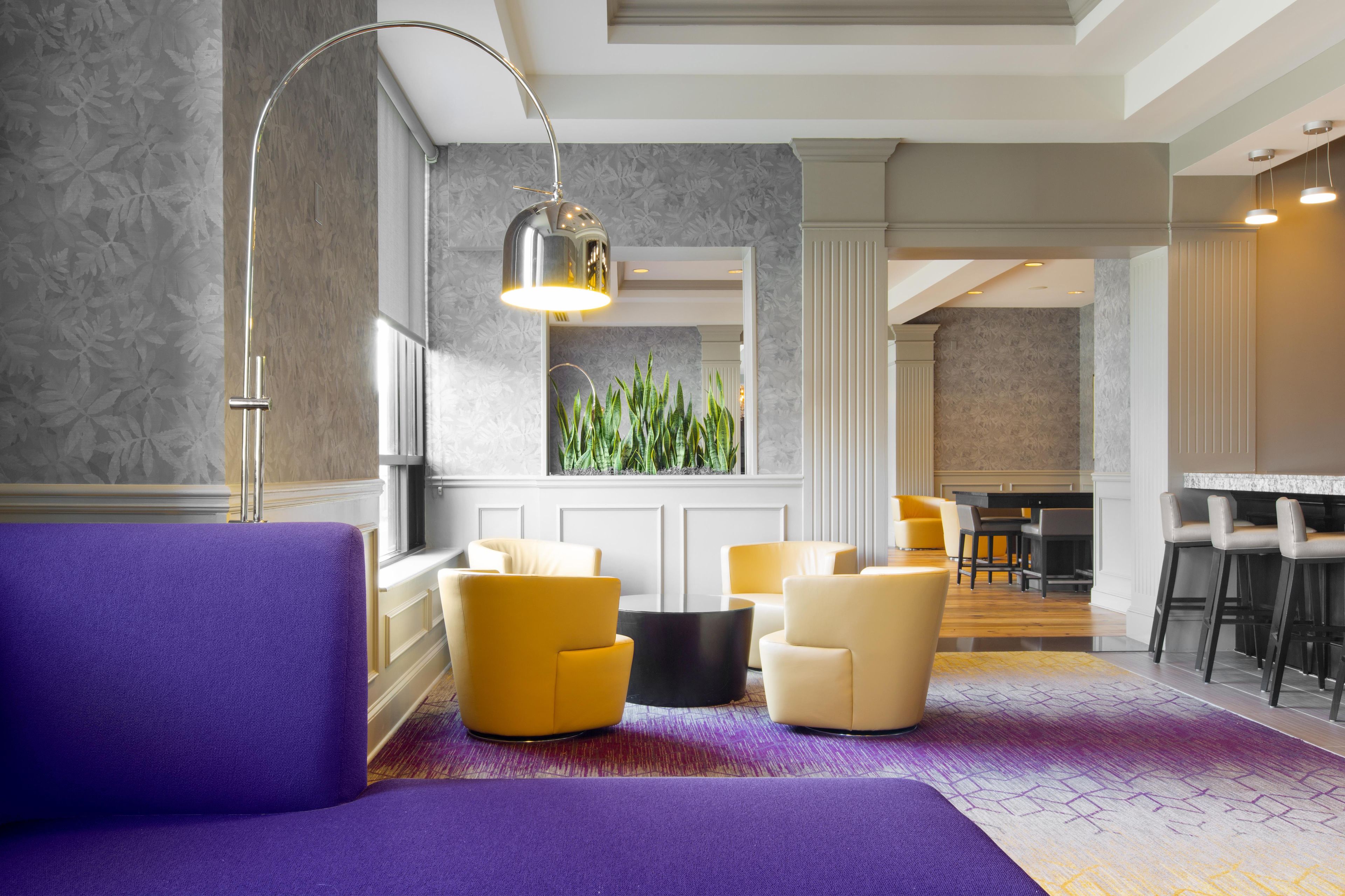 A modern hotel room with a purple sofa, yellow chairs, a black coffee table, and a large window with a view of greenery.