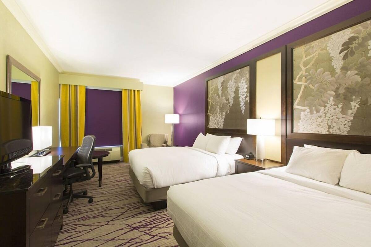 A hotel room with two beds, a desk, a chair, a TV, and a large window with curtains.