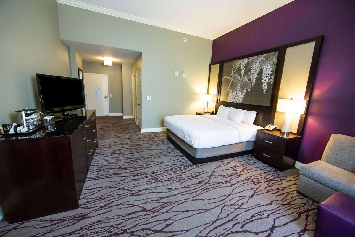 A hotel room with a bed, a desk with a flat-screen TV, a chair, a nightstand with a lamp, and a large mirror on the wall.