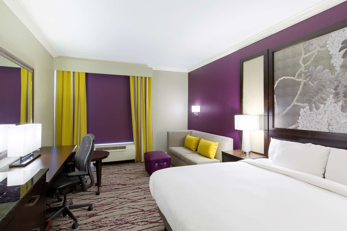 A hotel room with a bed, a desk with a chair, a sofa with yellow cushions, a purple ottoman, a nightstand with a lamp, and a large floral artwork on the wall.