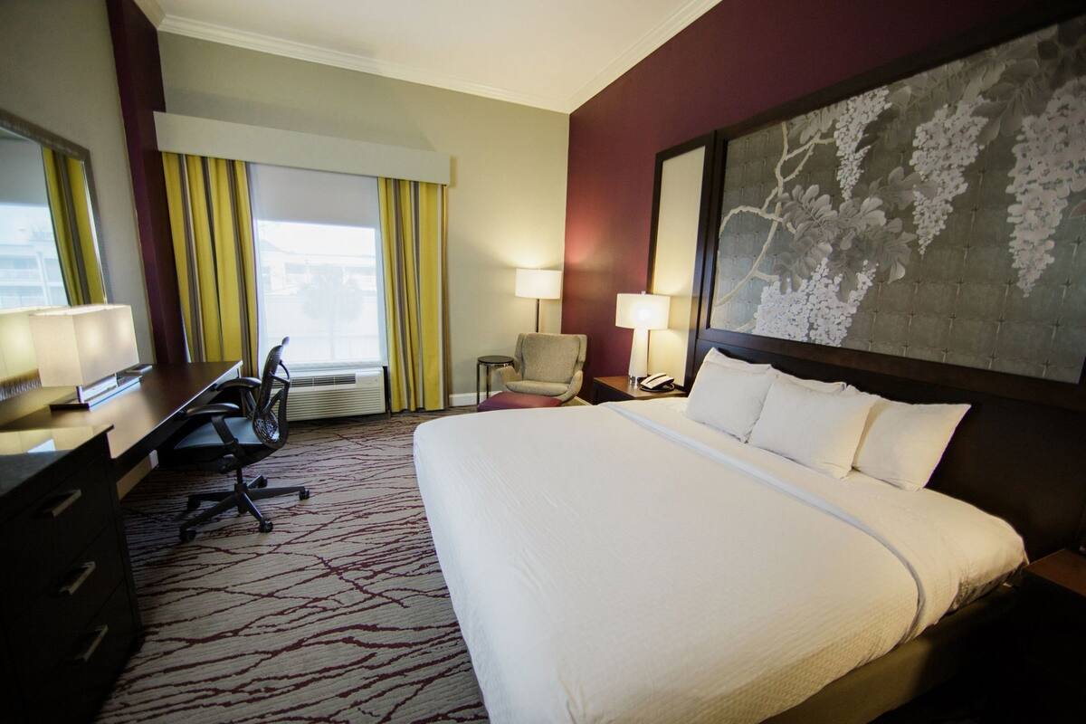 A hotel room with a large bed, a desk with a chair, a small table with a lamp, and a window with curtains.