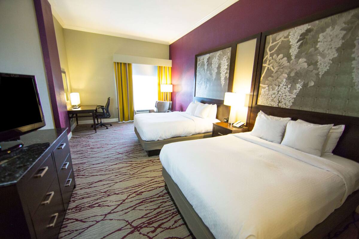 A hotel room with two beds, a desk with a chair, a television, and a large artwork on the wall.