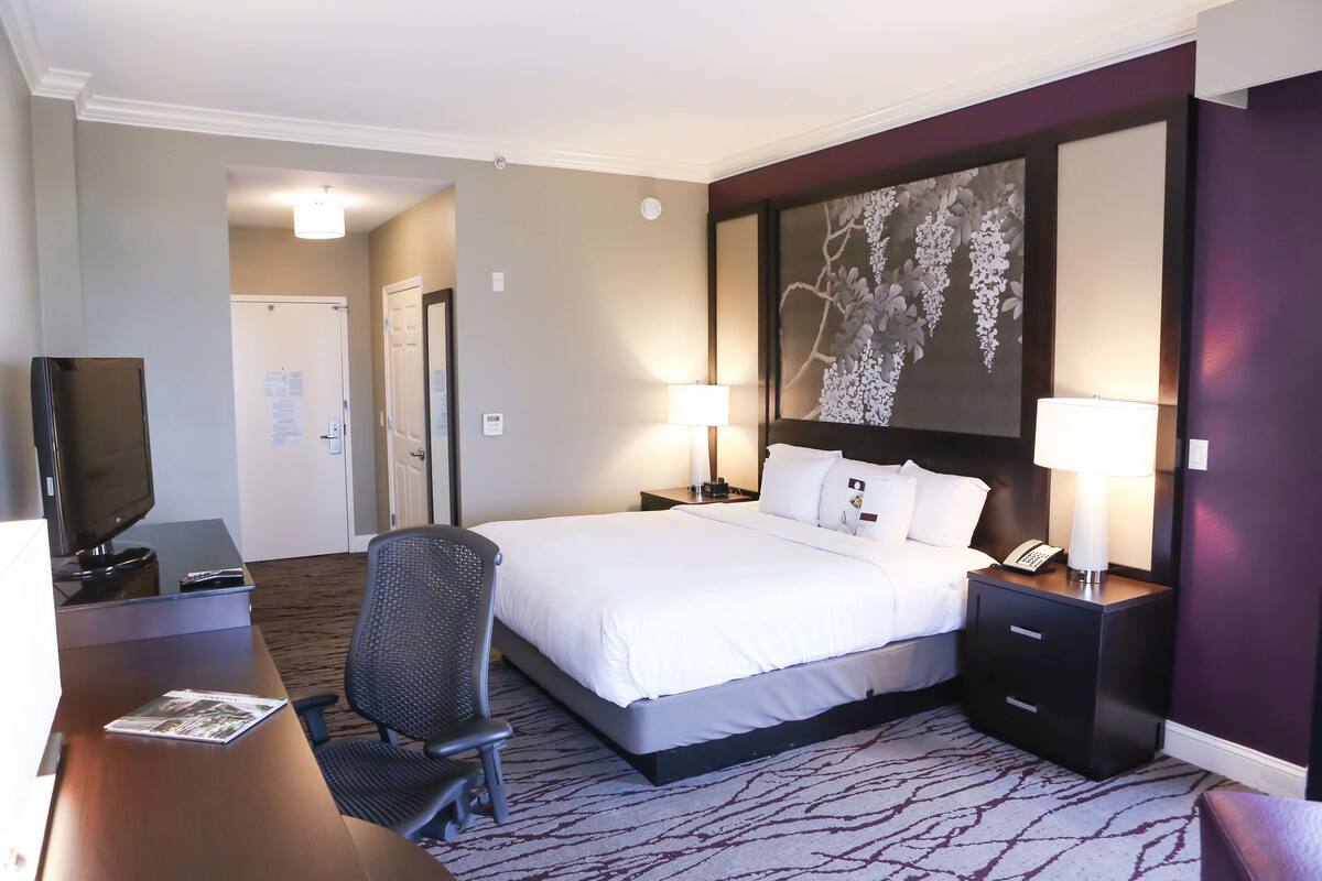 A hotel room with a bed, bedside tables, a desk with a chair, a television, and a large floral artwork on the wall.