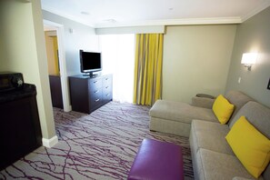 Habitación de hotel con sofá, televisión, una mesita, un armario oscuro y una alfombra estampada.