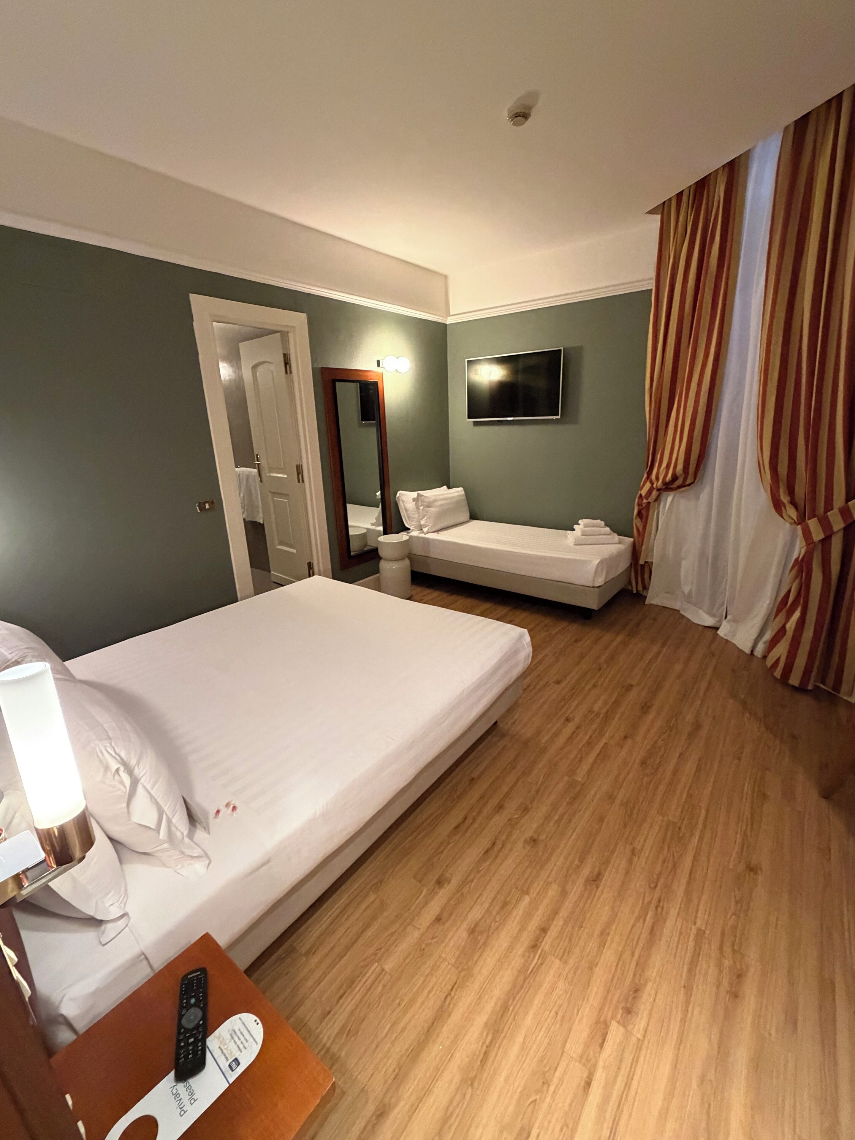A hotel room with a bed, a bedside table with a lamp, a TV mounted on the wall, and a bathroom visible through an open door.