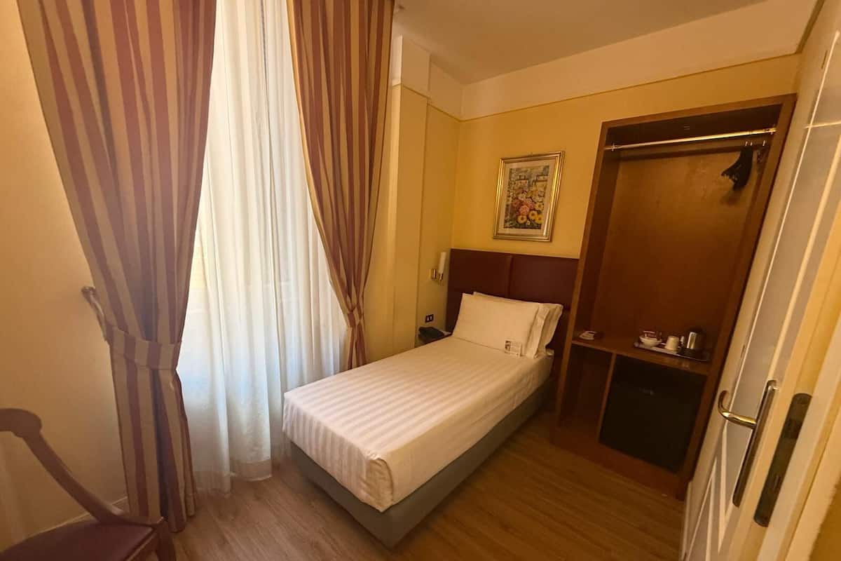 A small hotel room with a single bed, a wooden chair, a wardrobe, and a painting on the wall.